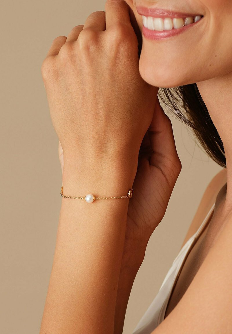 Bracciale in oro con un unico centro di perla bianca, indossato su una mano su uno sfondo neutro. Texture liscia con un design delicato.