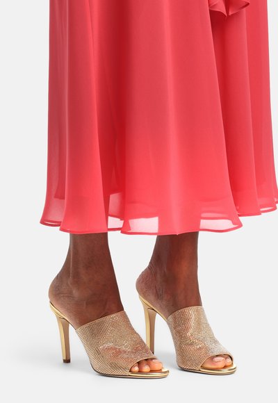 Pieds portant des mules dorées à talons hauts et bouts ouverts ornées de strass, associées à une robe midi corail fluide.