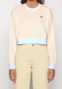 Felpa corta rosa chiaro con polsini e scollatura blu. Presenta un piccolo logo Adidas viola sul petto. Indossata con pantaloni di velluto a coste beige.