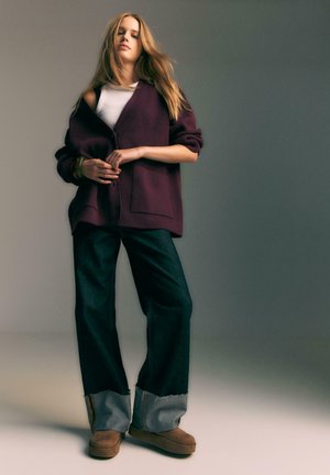 Übergroßer burgunderfarbener Cardigan mit vorderen Taschen, kombiniert mit weit geschnittenen, dunkelblauen Jeans mit ausgefransten grauen Säumen und braunen Stiefeln.