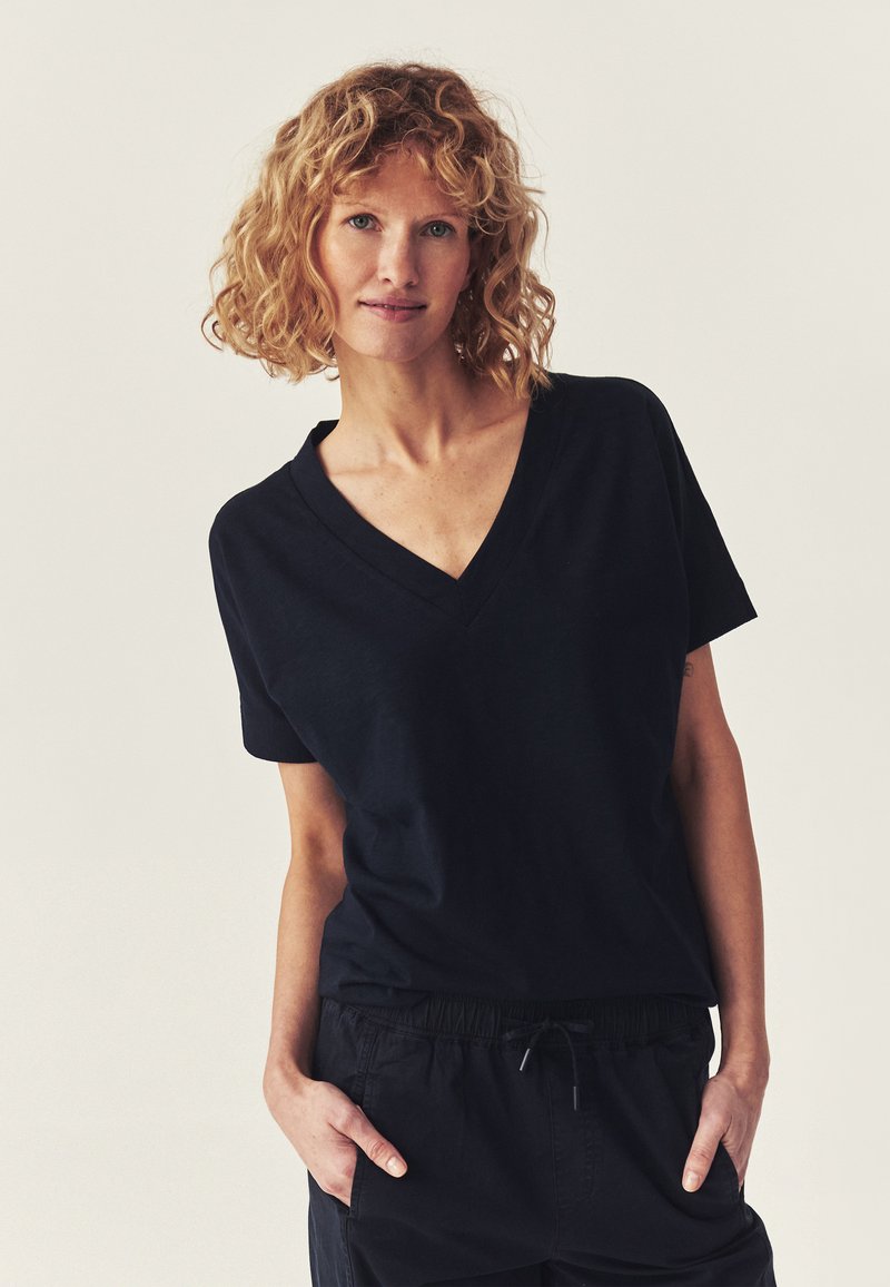 Femme aux cheveux blonds bouclés portant un t-shirt noir à col en V et un pantalon noir, les mains dans les poches, debout devant un fond clair uni.