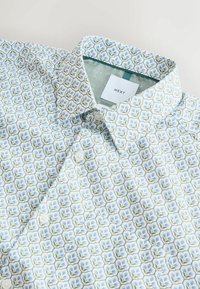 Chemise blanche avec un motif floral présentant des fleurs vertes et bleues. Tissu doux, col standard et patte de boutonnage à un seul bouton. Étiquette de la marque visible.