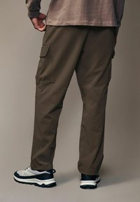 Pantalones cargo marrones con bolsillos laterales, corte recto, combinados con zapatillas negras y blancas, tejido texturizado y cintura elástica.