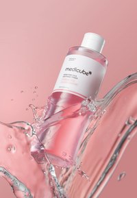 Bottiglia di toner rosa in plastica trasparente con tappo bianco, etichetta con testo minimale. Spruzzi d'acqua attorno alla bottiglia su uno sfondo rosa.