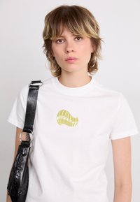 White cotton t-shirt featuring a green shell graphic. The shirt has a classic fit, short sleeves, and a round neckline. Black strap bag adjoined.