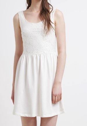 Femme portant une robe blanche sans manches avec un corsage en dentelle et une jupe fluide, debout devant un fond blanc uni.