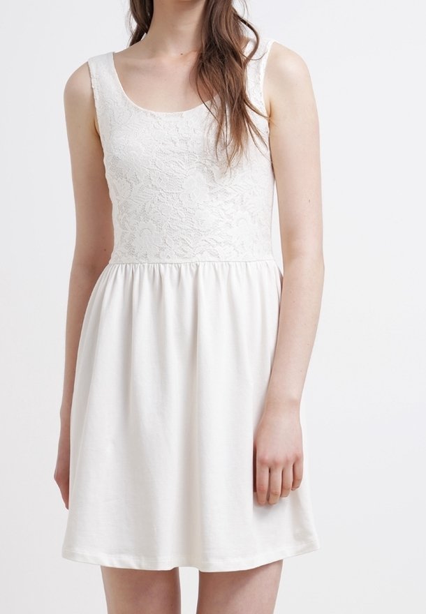 Femme portant une robe blanche sans manches avec un corsage en dentelle et une jupe fluide, debout devant un fond blanc uni.