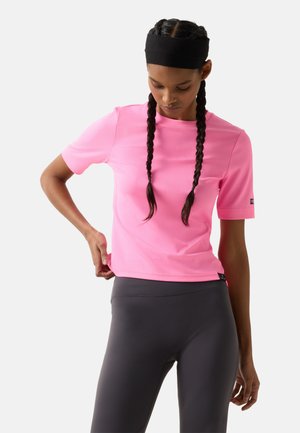 Vrouw met gevlochten haar en zwarte hoofdband draagt een felroze top met korte mouwen en donkergrijze legging, terwijl ze haar shirt rechtzet.