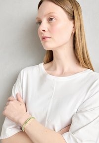 Valge puuvillane T-särk ümmarguse kaeluse jaotusega. Roheline helmestest käevõru randmel, millel on erinevad tekstuurid ja geomeetrilised kujundid.