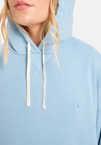 Ljusblå hoodie i mjukt tyg med dragsko och en liten broderad logotyp på bröstet. Texturerad detaljering vid halsen.