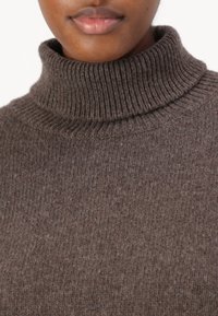 Καφέ μάλλινη πουλόβερ με ψηλό λαιμό, με ραβδωτή υφή και γιακά. Κοντινή λήψη που αναδεικνύει το σχέδιο πλέξης και την απαλή επιφάνεια του υφάσματος.