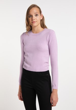 Jonge vrouw draagt een lichtpaarse trui en zwarte broek, met lang haar en een roze haarspeld, staand tegen een effen witte achtergrond.