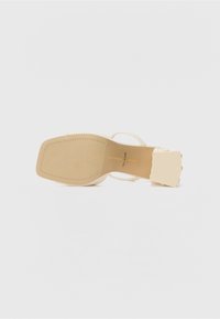 Sandale à talon beige avec une sangle texturée, bout carré et talon épais. Semelle lisse avec couture décorative et détail de logo.