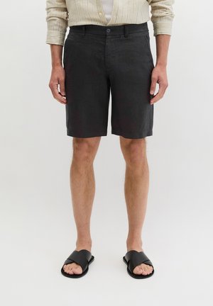 Mann trägt schwarze Shorts, beige gestreiftes Langarmshirt und schwarze Slide-Sandalen, steht auf weißem Hintergrund.