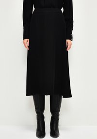 Jupe midi noire en tissu lisse, dotée d'une taille ajustée et d'une silhouette fluide, associée à des bottes noires montant jusqu'aux genoux.