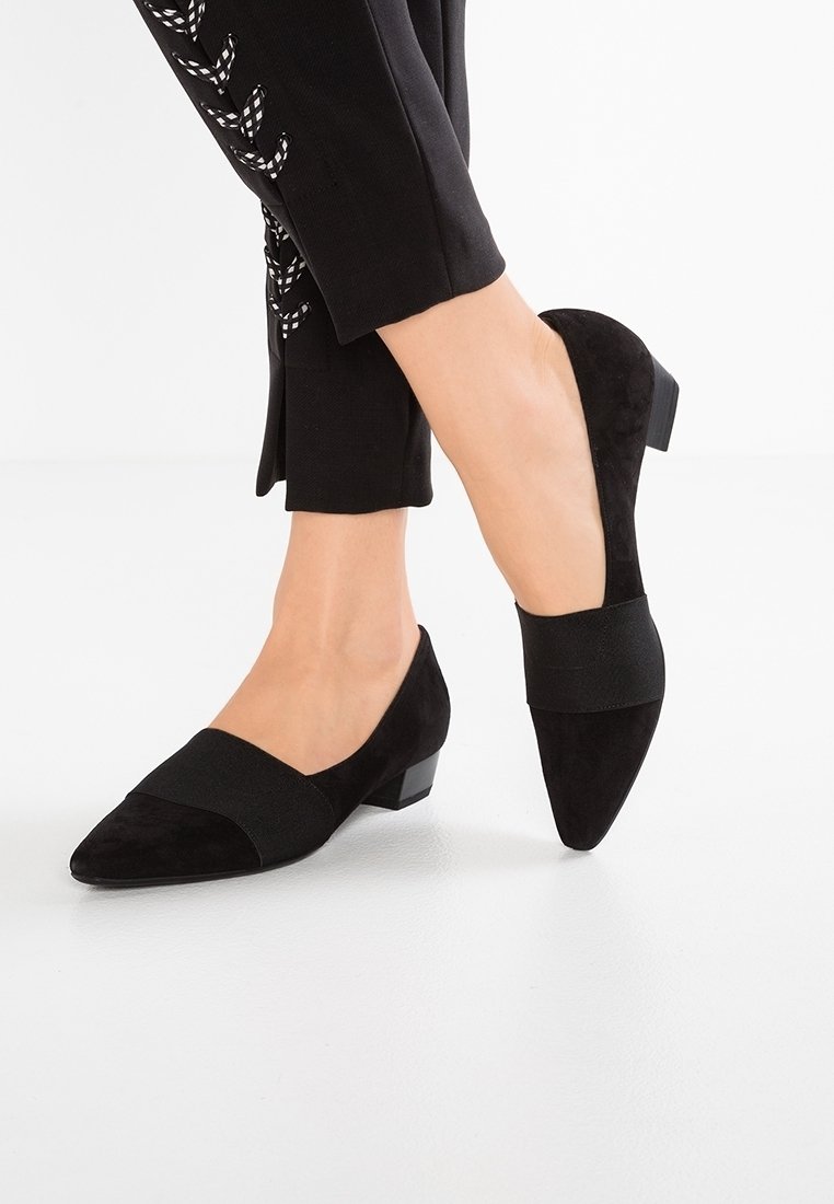 Femme portant des chaussures en daim noir à talon bas et bout pointu avec une large bande élastique, associées à un pantalon noir avec un détail lacé blanc.