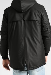 Man som bär en svart huva jacka med detaljer på bakstycket och ljusblå jeans, stående vänd bort från kameran.