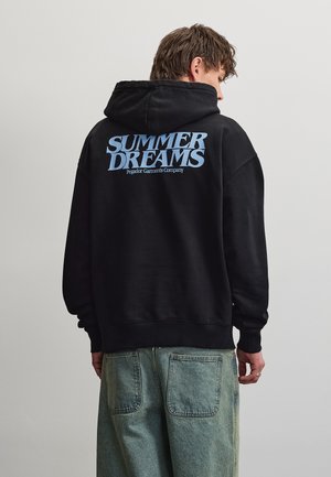 Ung mand iført sort hættetrøje med lyseblå tekst "SUMMER DREAMS Pegador Garments Company", kombineret med løse, falmede blå jeans, vendt væk.