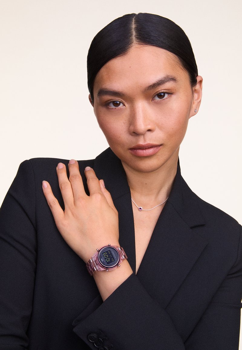 Montre numérique avec un cadran noir, un bracelet transparent rose, un boîtier rond et un affichage numérique. Portée au poignet sous un blazer noir.