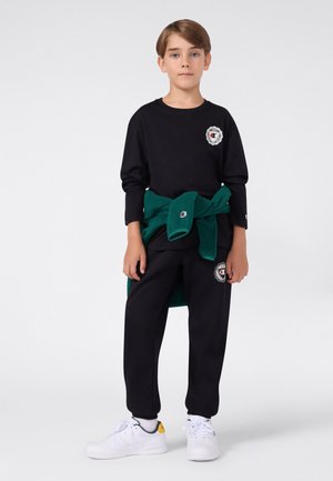 Ein Junge steht mit dem Gesicht nach vorne, trägt ein schwarzes Langarmshirt und Hose mit Logos, weiße Sneakers und einen grünen Pullover, der um die Taille gebunden ist.