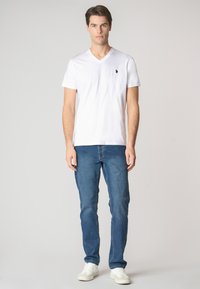 T-shirt bianca in cotone con scollatura a V e un piccolo logo nero sul petto, abbinata a jeans in denim azzurri a gamba dritta e sneaker bianche.