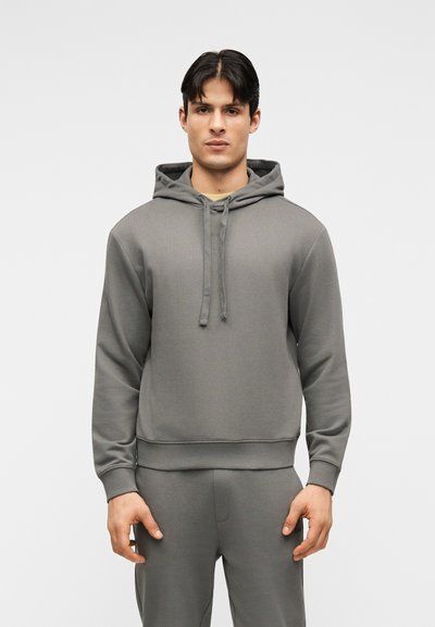 Homme debout portant un sweat à capuche gris assorti et un pantalon de jogging, les mains détendues le long du corps, sur un fond uni.