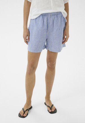 Vrouw draagt blauw-witte verticale gestreepte korte broek, wit topje en zwarte slippers, staand op een witte achtergrond.