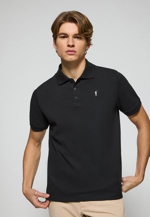 Polo negra de algodón con cuello clásico y tres botones. Presenta un pequeño logo blanco en el lado izquierdo del pecho. Textura suave.