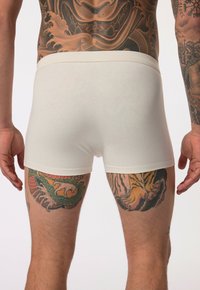 Boxers en coton blanc avec une texture douce, dotés d'une taille épaisse, portés par une personne avec des tatouages colorés sur les jambes.