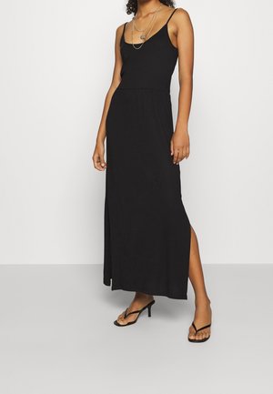 Femme portant une robe maxi noire sans manches avec une fente latérale, des colliers argentés superposés et des sandales à talons noires sur un sol gris et un fond blanc.