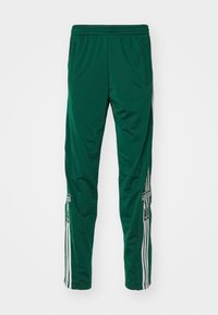 Zelené sportovní kalhoty vyrobené z lehkého materiálu, s bílými bočními pruhy, zipovými kapsami a logem adidas na přední straně.