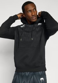 Μαύρο φούτερ με κουκούλα με υφή που θυμίζει κύματα, διαθέτει μπροστινή τσέπη και κορδόνια. Διακοσμητικά λογότυπα στο πάνω αριστερό μέρος του στήθους.