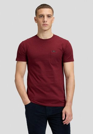 Mann steht vor einem schlichten Hintergrund, trägt ein eng anliegendes bordeauxrotes T-Shirt mit Brusttasche und dunkle Hose, schaut nach vorne.