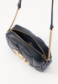 Borsa a tracolla nera in pelle trapuntata con accessori in oro, dotata di chiusura a zip e un logo decorativo in oro. Dettagli della catena e della tracolla in pelle visibili.
