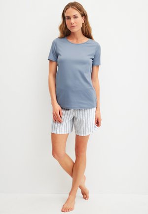 Woman standing barefoot against white wall, wearing light blue short-sleeve shirt and white shorts with blue vertical stripes.