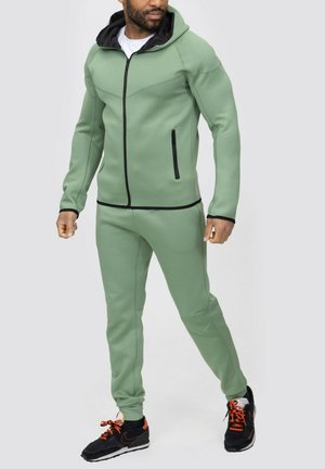 Survêtement de sport vert en tissu lisse, avec une veste à capuche zippée, des poches zippées sur les côtés et des accents noirs sur les zippers et le col.