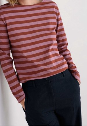 Person, die ein langärmliges rosa-braunes, horizontal gestreiftes Shirt in hoch taillierte schwarze Hose mit Händen in den Taschen trägt.