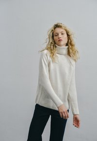 Sweter w kolorze kremowym z golfem, o luźnym kroju, z prążkowanymi mankietami i hemem oraz bocznym rozcięciem. Miękka tekstura, minimalistyczny styl.