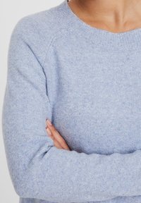 Jasnoniebieski sweter dzianinowy z okrągłym dekoltem, prążkowanymi mankietami i miękką teksturą. Materiał wydaje się przytulny i lekko zróżnicowany kolorystycznie.
