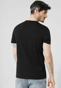 Černé tričko s krátkým rukávem zobrazené zezadu, s klasickým kulatým výstřihem, hladkou texturou a přiléhavým střihem. Viditelné tetování na paži.