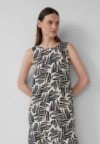 Robe sans manches avec un motif de feuilles noir et crème, fabriquée en tissu léger, avec un col rond et un ourlet en volants.
