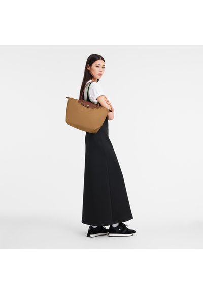 Brown tote bag with dual handles, featuring a front pocket and a structured design. Worn by a person in a long black dress and black sneakers.