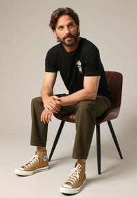 Zwarte t-shirt met een klein doodshoofd ontwerp, olijfgroene broek en tan hoge sneakers met witte veters, zittend op een bruin leren stoel.