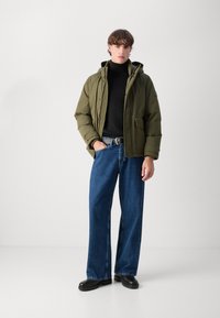 Grøn pufferjakke med hætte, sort turtleneck og bredbenede blå jeans. Sorte sko fuldender outfittet. Minimalistisk design.