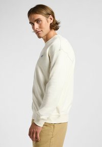 Sweatshirt de couleur crème avec un col rond, des poignets côtelés et un petit patch logo sur la poitrine ; texture douce et coupe standard.
