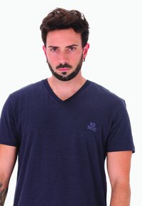 Uomo con capelli scuri e barba, vestito con una maglietta V-neck Diesel blu navy, in piedi contro uno sfondo bianco semplice.