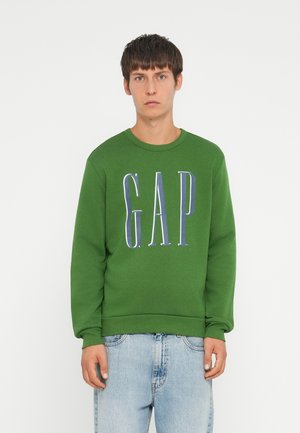 Green sweatshirt featuring front "GAP" logo in contrasting colors. Ribbed crew neck and cuffs, soft textured fabric. Worn with light blue jeans.