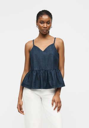 Femme aux cheveux tressés portant un top péplum bleu foncé sans manches et un pantalon blanc, debout devant un fond clair uni.