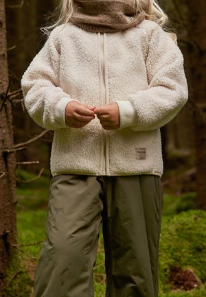 Bambino con giacca di pile beige e pantaloni verde oliva in piedi nella foresta, con le mani giunte davanti.