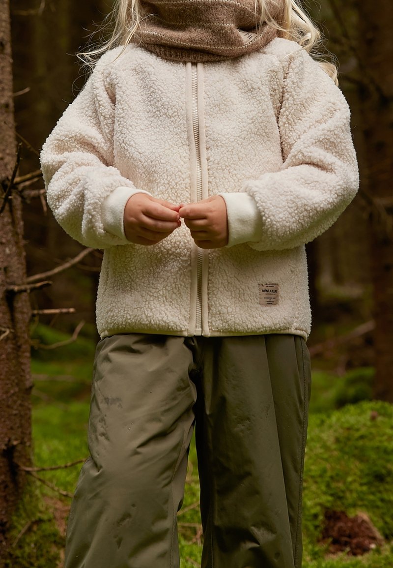 Enfant portant une veste en polaire beige et un pantalon vert olive debout dans la forêt, les mains jointes devant lui.